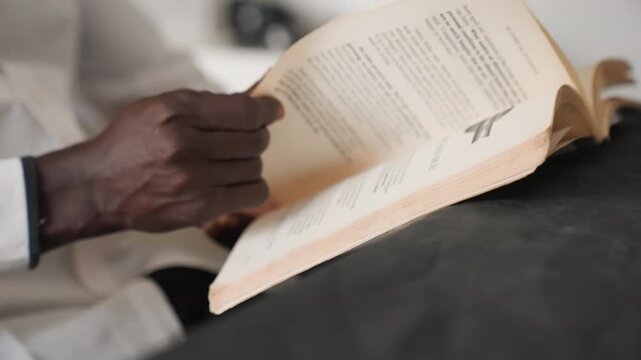 Hands turning pages of book closeup, gentle ambient light casting warm tones, paperback resting on lap, sleeve and wrist visible, slow deliberate motion, textured paper and soft fabric, calm intimate