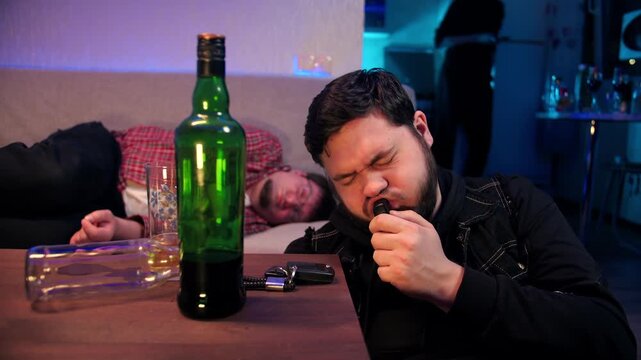 A drunk caucasian black-haired man blows into a breathalyzer to check his blood alcohol content. His friend sleeps on the couch in a cozy room. Bottles of alcohol and car keys are on the table