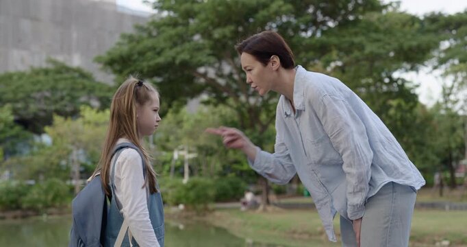 Portrait of worried mother scolds little daughter for bad rating in city park. Woman criticizes junior schoolgirl and threatens with finger on garden background
