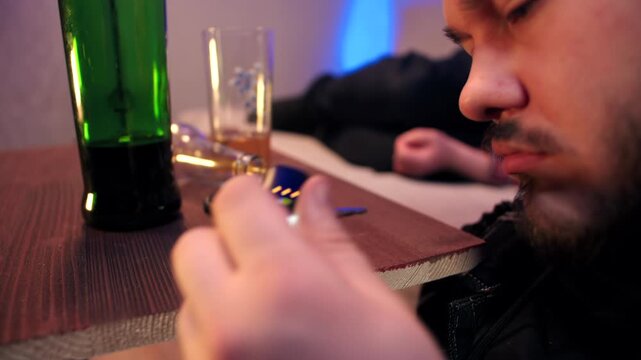 Close-up of a bearded caucasian man blowing air into a breathalyzer to check blood alcohol content. A bottle of whiskey and car keys are on table in apartment. Problem of alcoholism and drunk driving