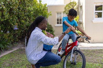 Naklejka premium African American man and child boy kneeling front yard steadying red bike, holding blue helmet