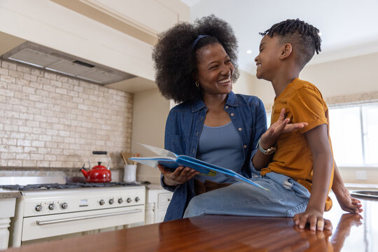African American mother and school-age son reading open picture book at wooden kitchen island
