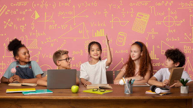 A group of five students sits at a table in a classroom. They engage in a discussion while sharing books and a laptop. One student raises a hand to answer a question, while others watch.