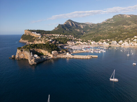 Aerial view of Port de Soller harbor with sailboats, the lighthouse on the rocky cliff, and the Tramuntana mountains under a clear sky Port de Soller, Balearic Islands, Spain.