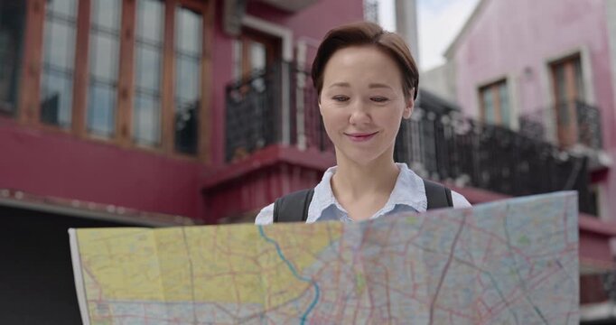 Portrait of cheerful lady tourist reads map as stands against old buildings. Short haired female with backpack studies town plan of landmarks in city center