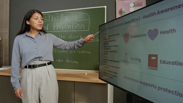 Medium shot of young Hispanic female teacher talking to school students about sexually transmitted infections, highlighting importance of awareness, prevention and testing, pointing to screen