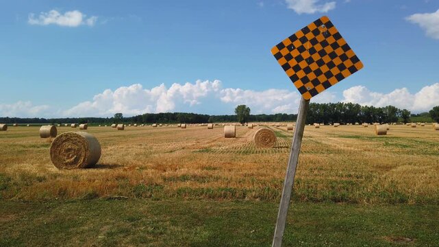 End of a road looking to a farmland field with rolled bales of hay livestock feed. A checkered road sign indicating a dead end caution.
