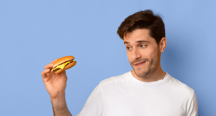 A man stands holding a burger in his right hand. He looks at the burger with curiosity and a slight smile. The background is plain blue. The man wears a white t-shirt. © Prostock-studio