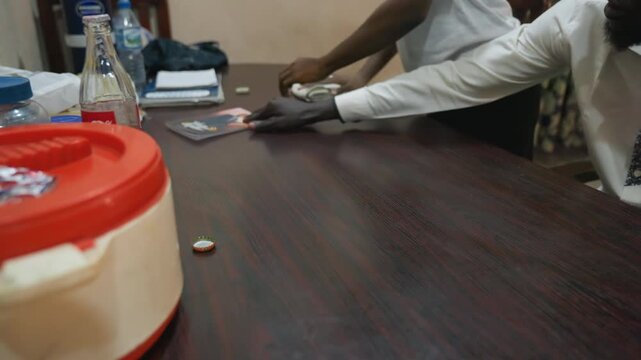 Black man cleaning office table with coworkers reaching across, wiping surface with cloth near plastic container and bottle, casual shirts and sleeves visible, hands moving documents and magazine,