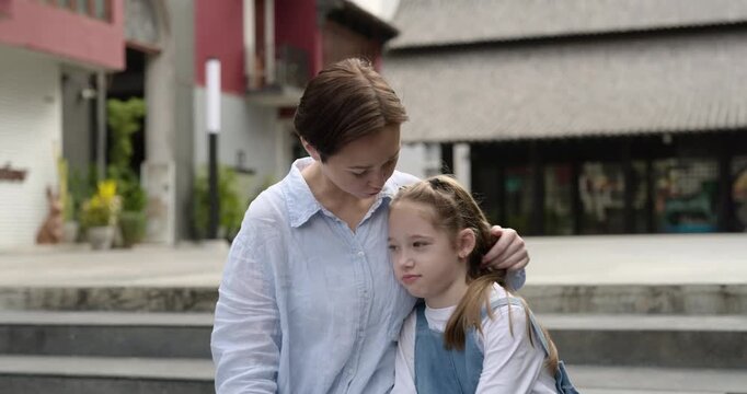 Portrait of supportive mother calms down sad little daughter while sits on parapet. Woman embraces girl feeling bad as rests on town street. Loving family