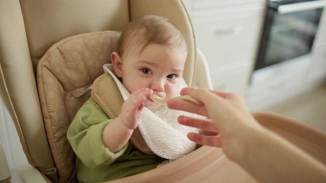 A baby sits in a high chair in the kitchen and is spoon-fed porridge. The baby eats and grimaces as he feeds