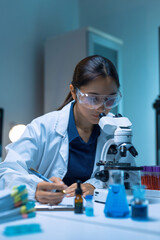 Female scientist wearing safety goggles, looking through microscope and writing down observations, conducting research in a lab © Crystal