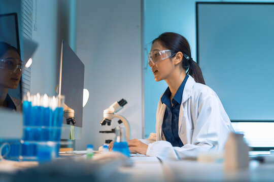 Female scientist wearing safety glasses and lab coat, focusing on computer screen while conducting research in a modern lab
