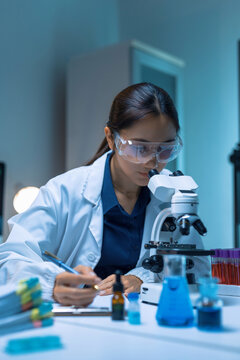 Female scientist wearing safety goggles, looking through microscope and writing down observations, conducting research in a lab