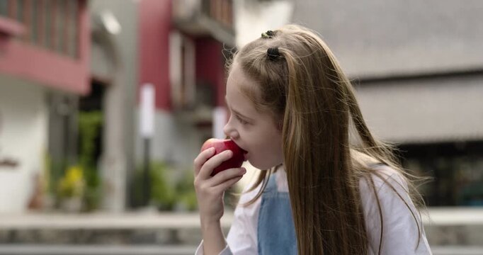 Portrait of little girl with pigtails eats red apple as stands on city street. Cute junior schoolgirl bites piece of fresh fruit in old district. Healthy snack