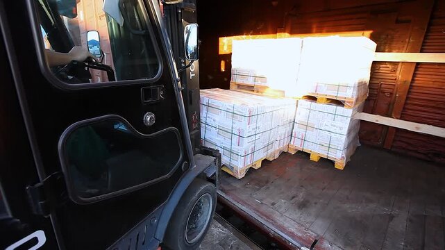 A forklift is loading pallets of products into a covered freight car. Loading a freight car in the factory yard. A freight handler loads a railcar with boxed products. Loading finished products