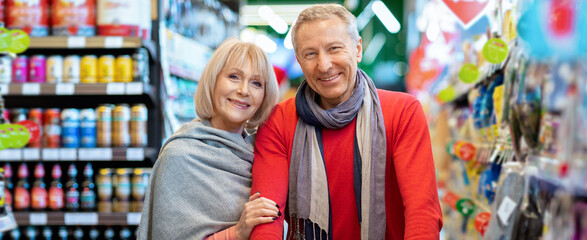 A couple stands close together in a grocery store, smiling while looking at the camera. They hold onto a shopping cart and are surrounded by shelves filled with various products. © Prostock-studio