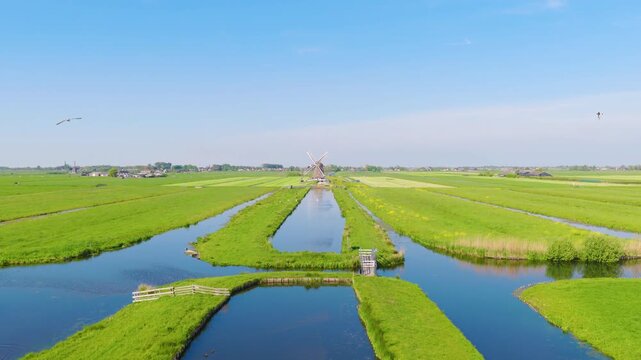 Aerial view of traditional Dutch windmills near water canals in a green field. Clear sunny day with reflections on the water surface and a wide video landscape captured by drone.