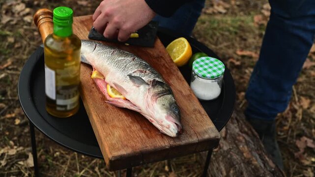 Close up of male hands stuffing fish with lemon slices before cooking over a campfire. Outdoor food preparation scene in the forest with camping atmosphere.