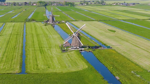 Aerial view of traditional Dutch windmills near water canals in a green field. Clear sunny day with reflections on the water surface and a wide video landscape captured by drone.