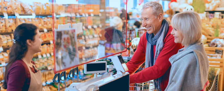 Two customers are at a grocery store checkout talking to a cashier. They are smiling and sharing a light moment while surrounded by various products on display.