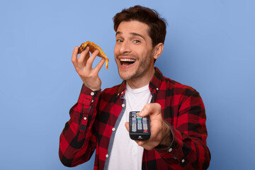 Naklejka premium A man enjoys a snack while watching TV. He holds a remote in one hand and food in the other. He is standing in front of a plain blue background.