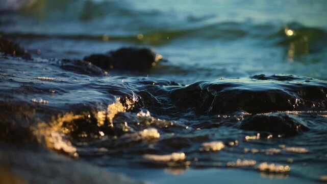 Close-up of water splashing over dark rocks at the shoreline. The scene captures the movement of waves and the texture of the stones.