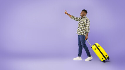 A man stands on a purple background holding a yellow suitcase. He raises one hand and points ahead, looking ready for travel or an adventure. The moment captures excitement and anticipation. © Prostock-studio