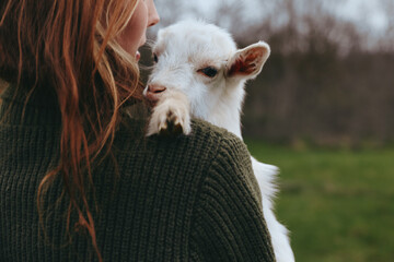 Obraz premium Goat kid rests against a smiling woman outdoors, gentle moment in soft light, warm knit sweater, rustic field backdrop, candid lifestyle scene, natural colors, peaceful companionship, countryside