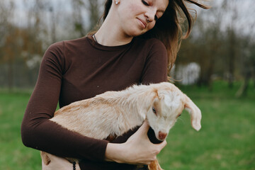 Obraz premium Lifestyle moment with a woman outdoors, gently holding a goat kid, warm sunlight, casual sweater, soft breeze, grassy field, natural colors, peaceful bond and connection