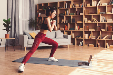 A woman in workout clothes performs lunges on a mat at home. She focuses on her exercise while a laptop sits in front of her. The room is bright and features bookshelves and plants. © Prostock-studio