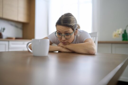 Tired woman staring at coffee mug at kitchen table