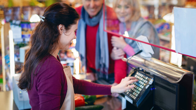 A worker at a grocery store scans items for customers at the register. Two shoppers stand nearby, waiting for their turn. The setting shows a busy store with various products around.