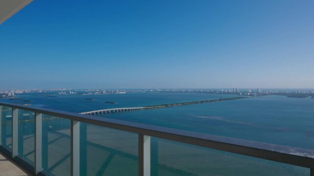 The balcony view shows calm water stretching across scene, distant skyline rises with tall buildings, clear blue sky makes space bright, glass railing frames open