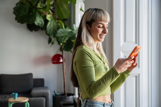 Smiling woman using smartphone at home near window light