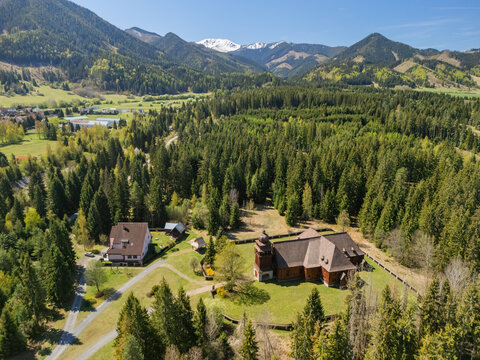 Aerial view of the Wooden Articular Church of Svaty Kriz surrounded by pine forests and rolling green hills with snow-capped mountains Svaty Kriz, Zilinsky kraj, Slovakia.