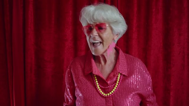 Funky grandmother with white hair wearing a shiny pink shirt, sunglasses, and extravagant jewelry dancing energetically against a red curtain background at a nightclub party