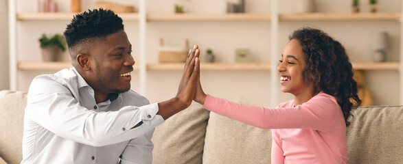 Fototapeta premium A father and his daughter are sitting on a sofa in their living room. They are smiling and playing a hand game. Sunlight comes through the window, creating a warm atmosphere.
