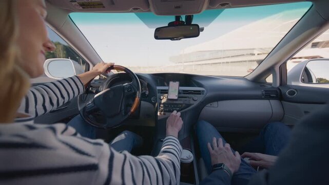 Caucasian woman driving car using GPS navigation on smartphone while passenger sits beside. Female driver holding steering wheel and adjusting map in daylight. Rear. Learning to drive. Back view.