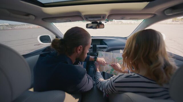 Man with tied hair driving car while female passenger holds paper map and points at location. Couple navigating route together inside vehicle during road trip in daylight through open parking area.
