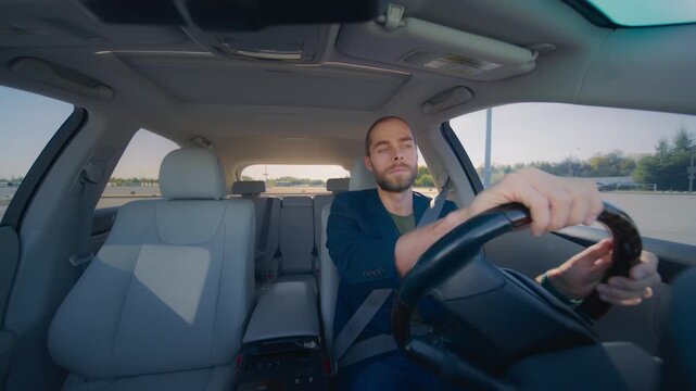 Caucasian man with brown hair in bun driving car, holding steering wheel with both hands, turning head to right. Bright daylight, empty passenger seat, road and trees outside. Male driver.