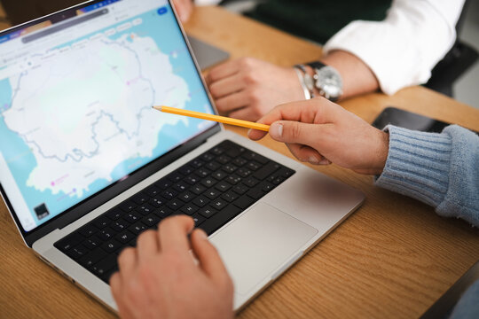 Two people collaborate on a laptop displaying a map, with one person pointing to the screen with a pencil, suggesting planning or research.