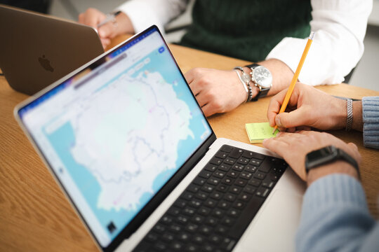 Two people collaborate at a desk, one using a laptop displaying a map, the other writing notes on a sticky pad.