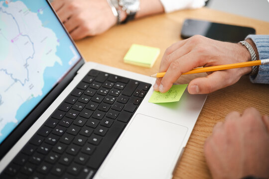 A person's hand holds a pencil over a sticky note on a laptop, planning a trip or project.
