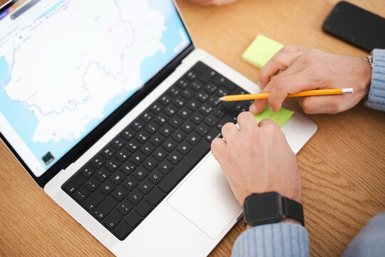 A person uses a laptop with a map displayed on the screen, holding a pencil over a sticky note.