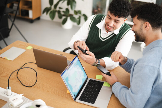 Two young men collaborate on a project, using laptops and smartphones to share information and discuss ideas at a wooden desk.