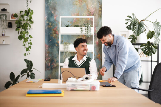 Two men collaborate at a desk in a bright, modern office space, discussing a project on a laptop.