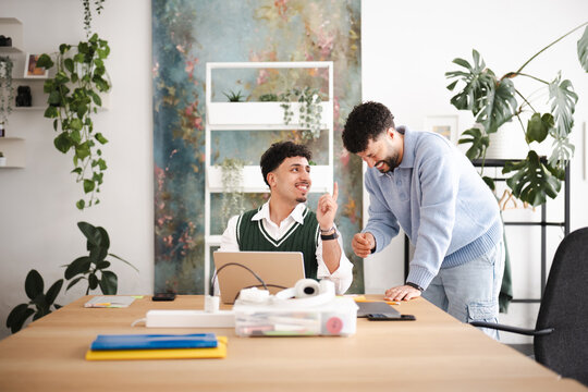 Two men collaborate at a desk with a laptop, one pointing and explaining an idea to the other who is smiling and giving a thumbs up.