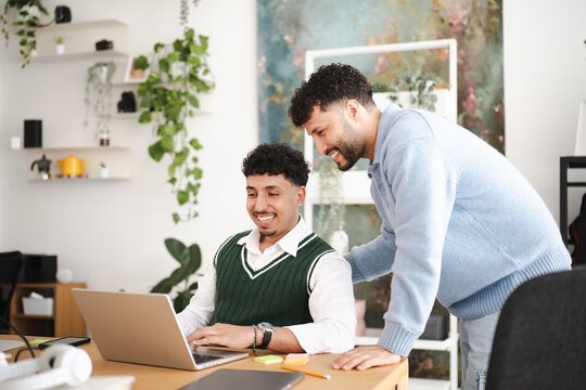 Two men collaborate on a laptop at a desk, one typing while the other leans in to observe and discuss.