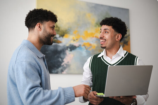 Two young men are collaborating on a project, one holding a laptop and the other pointing at the screen, both smiling and engaged in discussion.